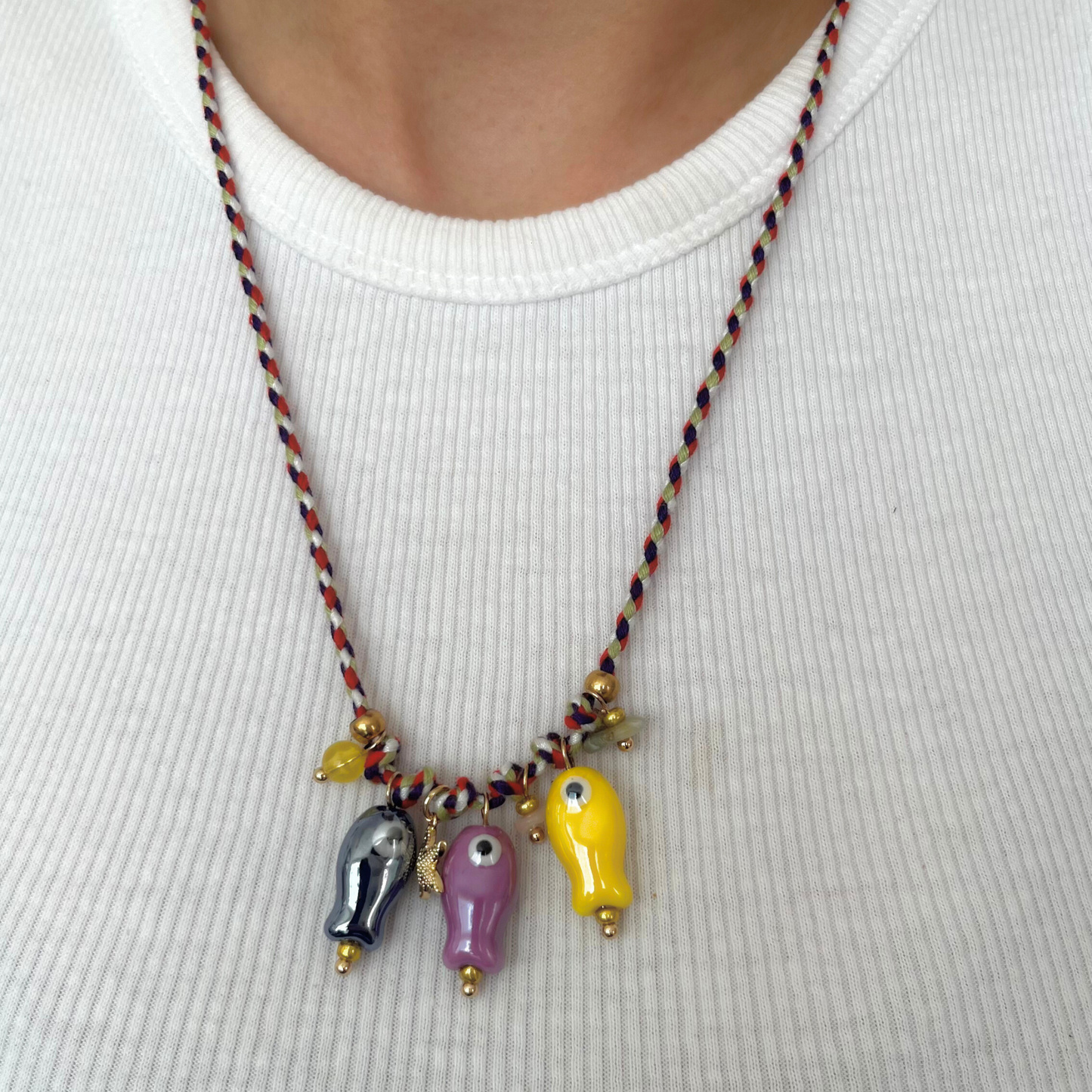 Collar artesanal con cordón trenzado y cinco dijes de peces de vidrio pintados a mano en tonos morado, rosa, amarillo y azul, sobre fondo claro.
