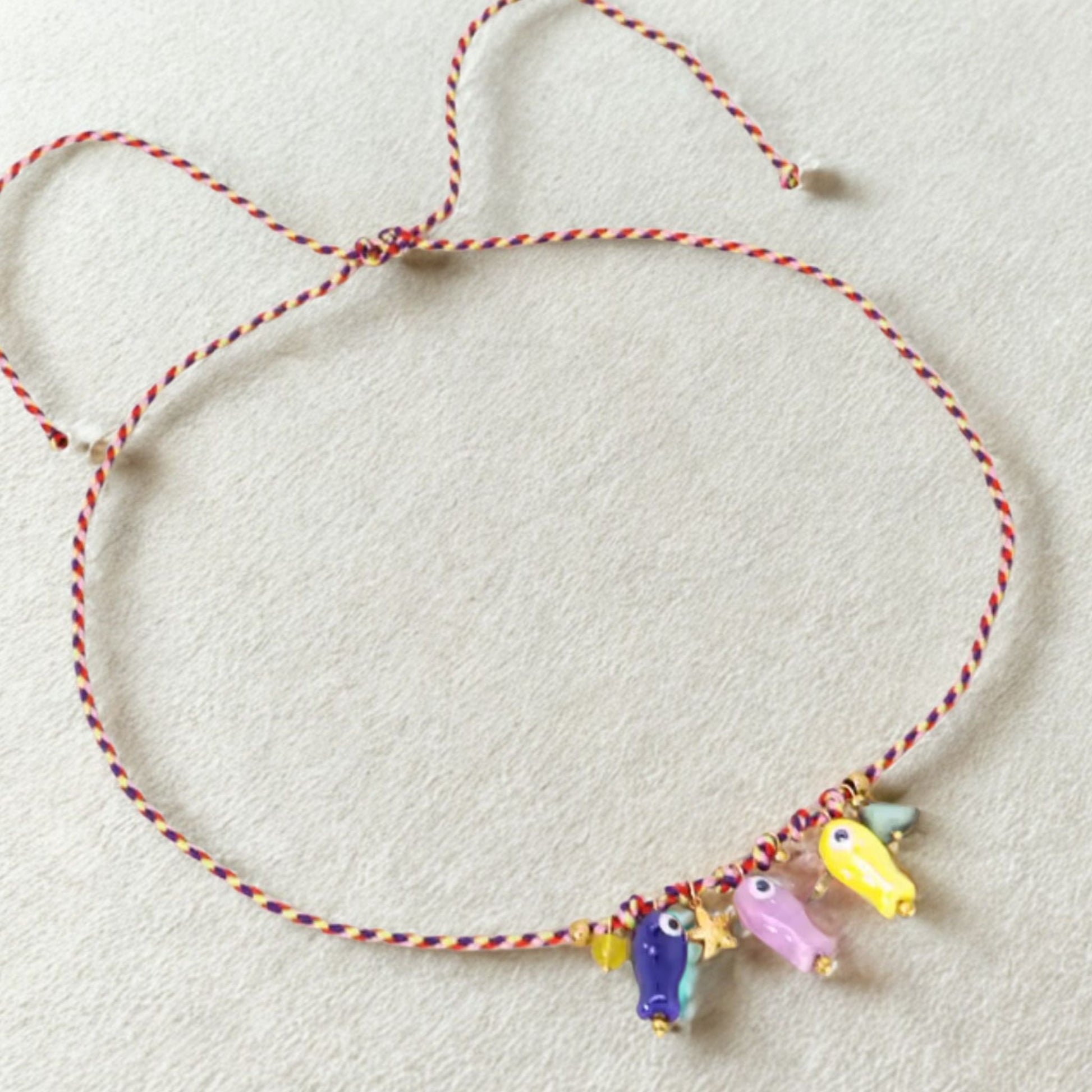 Collar artesanal con cordón trenzado y cinco dijes de peces de vidrio pintados a mano en tonos morado, rosa, amarillo y azul, sobre fondo claro.