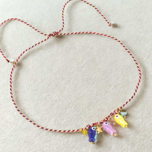 Collar artesanal con cordón trenzado y cinco dijes de peces de vidrio pintados a mano en tonos morado, rosa, amarillo y azul, sobre fondo claro.