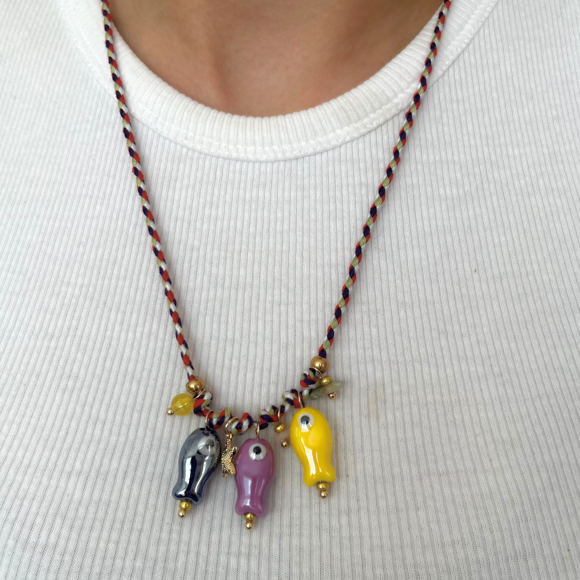 Collar artesanal con cordón trenzado y cinco dijes de peces de vidrio pintados a mano en tonos morado, rosa, amarillo y azul, sobre fondo claro.